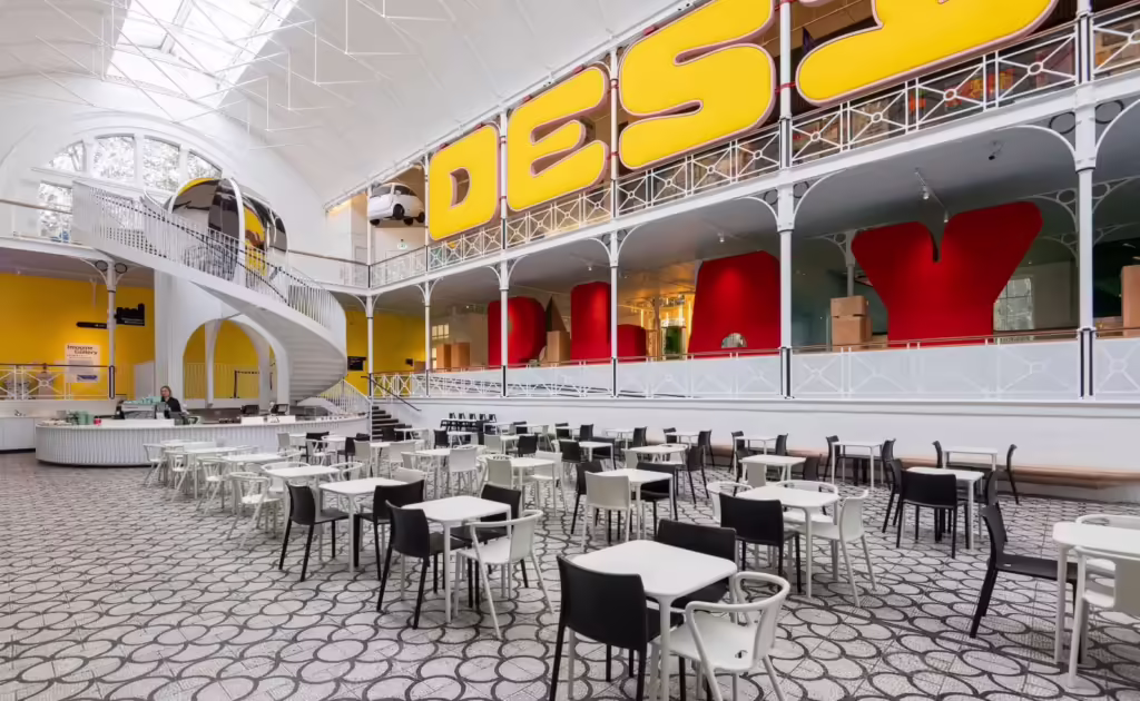 Pictured is the Young V&A cafe set up with white tables and black and white chairs. The cafe bar can be seen in the distance and there is a spiral staircase behind it leading up to the second floor.